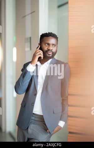 Content confident young Black businessman in jacket standing at wall and talking by mobile phone Stock Photo