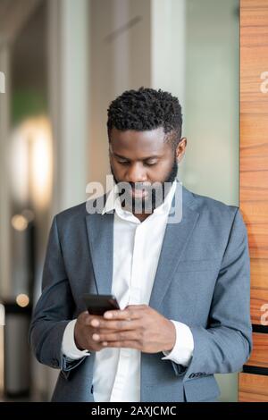 Serious young Black businessman in suit standing in corridor and using smartphone while texting with colleagues Stock Photo