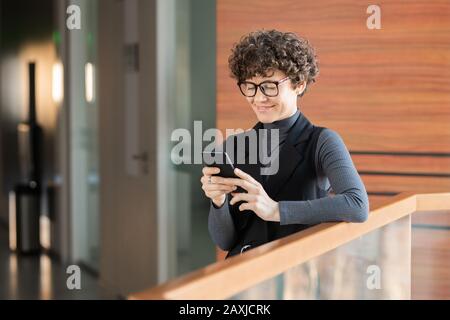 Smiling young curly-haired lady standing in corridor and checking social media statistics on phone Stock Photo