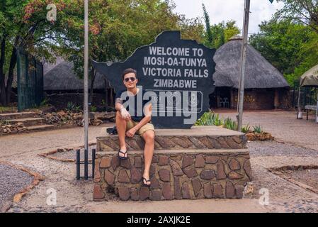 Tourist sits on the welcome sign placed at the entry of Victoria Falls, Zimbabwe Stock Photo
