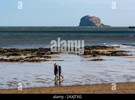 Milsey Bay, North Berwick, East Lothian, Scotland, United Kingdom. 12th Feb, 2020. UK Weather: A cold and windy but sunny morning on the beach at low tide with the Bass Rock volcanic plug on the horizon. The sea is choppy from the wind over the last few days. Two people walk on the sandy beach wrapped up in Winter clothing Stock Photo