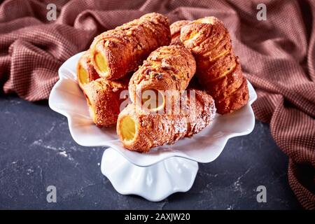 European traditional dessert: chimney cake or spit cake sprinkled with sugar and cinnamon served on a white cake stand on a dark background, close-up Stock Photo