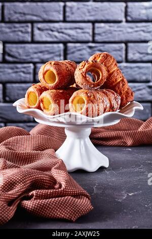 Cinnamon chimney cake or kurtos kalacs served on a white cake stand on dark concrete background in front of a black brick wall, close-up, vertical ori Stock Photo