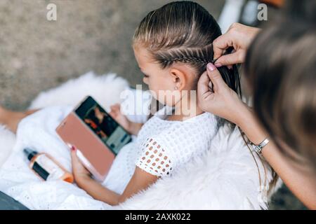 Mother does hair braid to her daughter, the girl is watching a cartoon on a mobile phone. Close up photo. Stock Photo