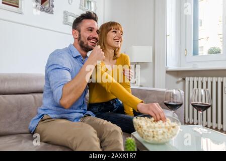 Happy beautiful couple having fun watching TV and eating pop corn at home sitting on the sofa. Stock Photo