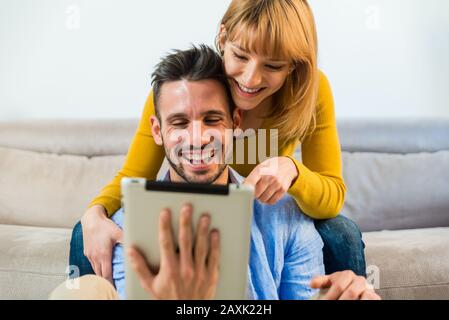 Happy couple of millennials using digital tablet at home. Boyfriend and girlfriend sitting on the sofa in the living room searching something on inter Stock Photo