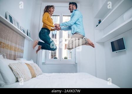 Happy couple in love having fun jumping on bed at home. Handsome man and beautiful woman are enjoying spending time together. Stock Photo