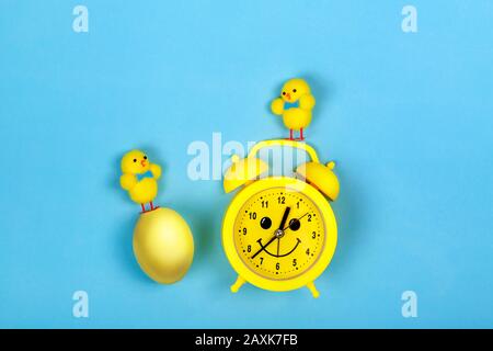 Retro yellow alarm clock with drawn smiling face and toys chicken and colored egg on blue background. Easter concept. Stock Photo