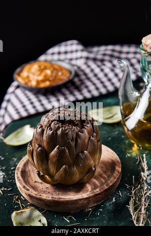 closeup of a roasted artichoke in a wooden plate, next to a cruet with olive oil and a plate with catalan romesco sauce in the background, on a dark g Stock Photo