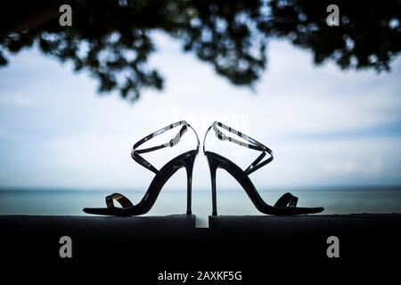 Close up of pair of shiny stiletto sandals, tree foliage and ocean in background. Stock Photo