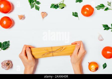 Uncooked spaghetti in woman's hands next to ingredients for making homemade pasta, tomatoes, garlic and parsley leaves, flat lay. Stock Photo