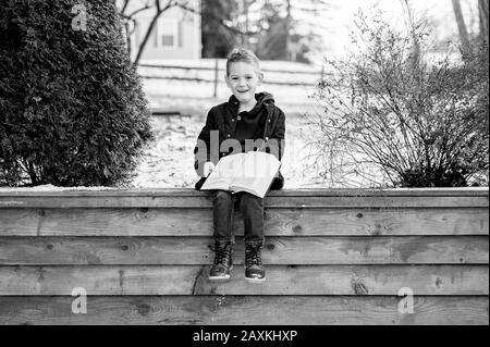 Grayscale of a happy boy sitting on a wooden fence and reading a book in a park Stock Photo