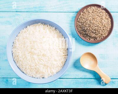 Long grain Basmati rice in ceramic bowl, cumin (jeera) in clay plate on blue wooden background Stock Photo