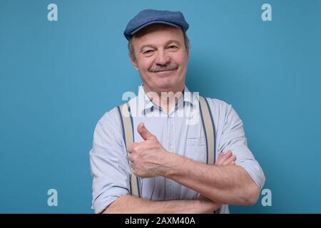 Portrait of friendly confident senior man in summer hat showing thumb up approving your choice standing isolated over blue background. Positive facial Stock Photo