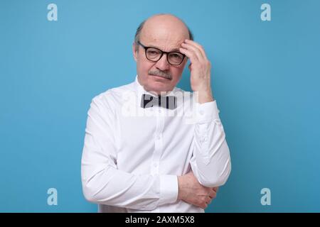 Senior man in glasses thinking and looking down trying to make a right solution. Studio shot on blue wall Stock Photo