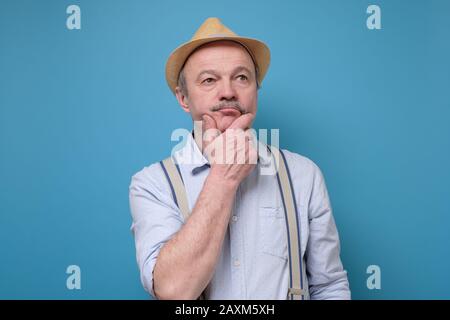 Senior man in summer hat thinking and rubbing a chin trying to make a right solution. Studio shot on blue wall Stock Photo