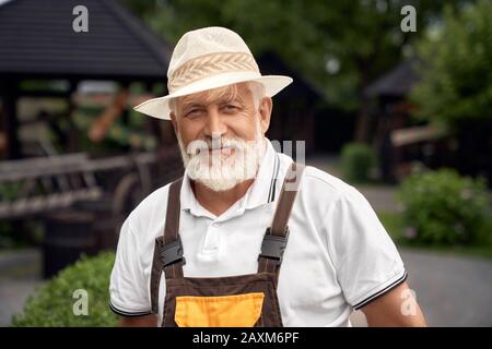 Portrait of positive senior man with grey beard wearing uniform and summer hat. Selective focus of eldery worker posing in garden near gazebos, looking at camera. Concept of gardening. Stock Photo