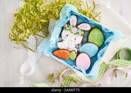 From above view of glazed cookies in shape of lovely bunnies, three eggs and carrot lying in box with blue napkin, ribbons isolated on white background. Spring and easter holidays concept. Stock Photo