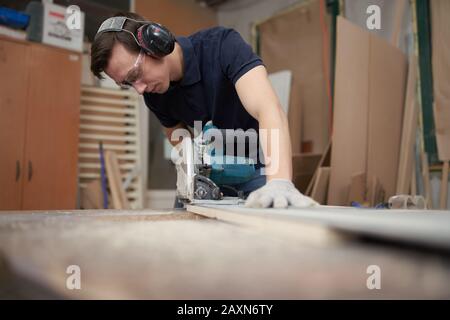 Carpenter man in protective headphones and white gloves works on jigsaw in workshop, close-up Stock Photo