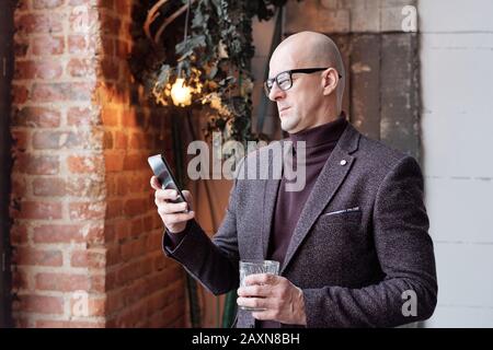 Serious bald man in turtleneck and jacket standing in lobby and checking message on phone Stock Photo