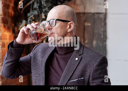 Rich middle-aged bald man in glasses standing in loft restaurant and drinking alcohol from glass Stock Photo