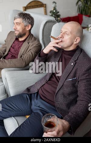 Relaxed bald businessman in fashionable outfit sitting in armchair and smoking cigar with friend Stock Photo