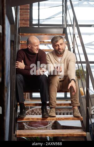 Handsome bearded man with whisky glass sitting on stairs and sharing life story with friend Stock Photo