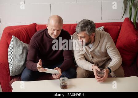 Above view of bald man sitting on sofa and using tablet while discussing internet news with friend Stock Photo