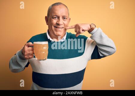 Senior handsome man drinking jar of beer standing over isolated yellow background smiling cheerful showing and pointing with fingers teeth and mouth. Stock Photo