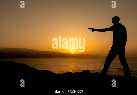 Silhouette of man holding gun at sunset near ocean.