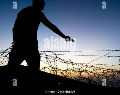Silhouette of man pointing gun near barbed wire fence.