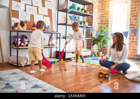 Beautiful teacher and toddlers playing tennis using racket around lots of toys at kindergarten Stock Photo