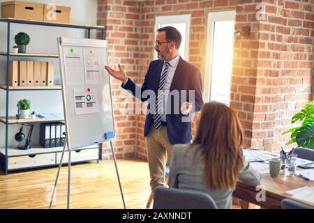 Two middle age business workers smiling happy and confidentt working together with smile on face. Man standing doing work presentation using board and Stock Photo