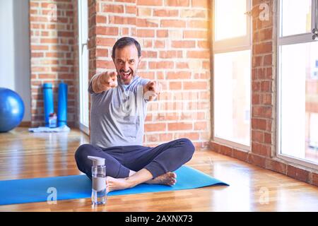 Middle age handsome sportman sitting on mat doing stretching yoga exercise at gym pointing to you and the camera with fingers, smiling positive and ch Stock Photo