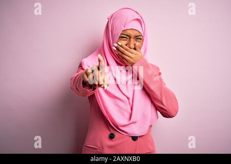 Young African American afro woman wearing muslim hijab over isolated pink background laughing at you, pointing finger to the camera with hand over mou Stock Photo