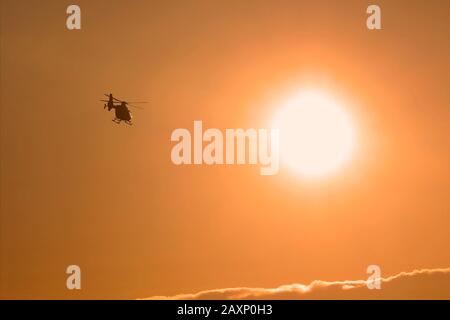 Helicopter in silhouette flying towards sunset against orange sky. Stock Photo