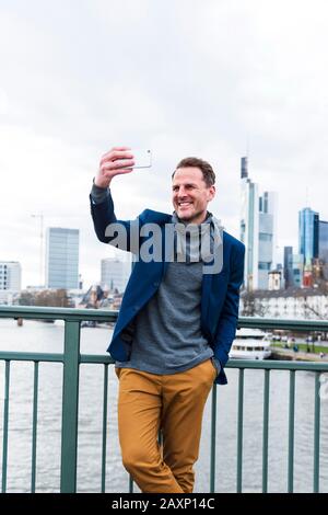 Businessman takes a photo of himself with Smartphone in front of the skyline at the Main, Frankfurt on the Main, Hessen, Germany Stock Photo