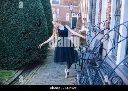 Young ballerina woman with red hair in ballet costume and pointe shoes is in beautiful pose dancing on street. Ballerina is posing in the streets. Gir Stock Photo