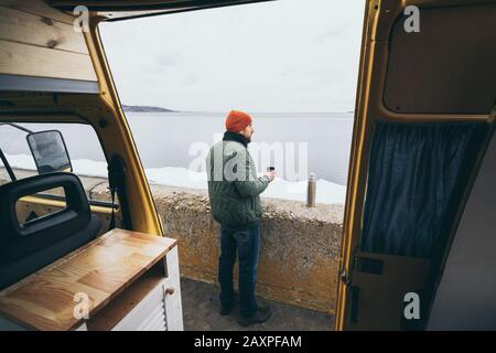 Young man having a hot drink from thermos in front of camper van overlooking a lake on background Stock Photo