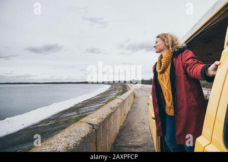 Young blond woman looking out of camper van overlooking the frozen winter sea Stock Photo