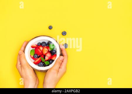 Woman hands holding coconut bowl with fresh ripe berries on yellow background. Top view with copy space Stock Photo