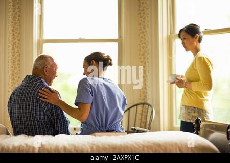 Female nurse puts a reassuring hand on the back of a senior man as they sit on the side of a bed while a woman stands with a cup of tea watching them solemnly. Stock Photo