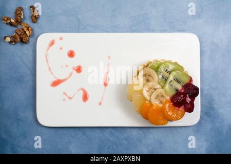 Cookie dough bowl with various kinds of fruit covered in jelly, on a white plate with syrup spilled over it. Selective focus on the cookie. Stock Photo