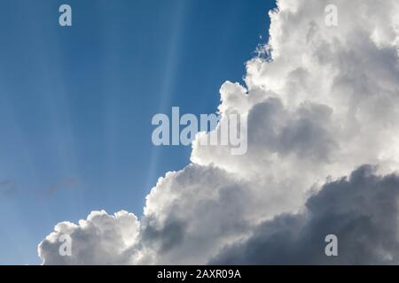 Sun rays shining from behind white clouds against a blue sky Stock Photo