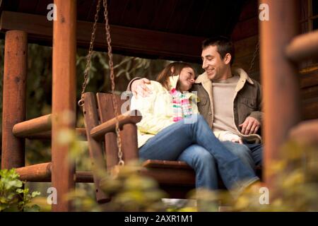 Young couple sitting on a hammock chair on the stoop of a log cabin. Stock Photo
