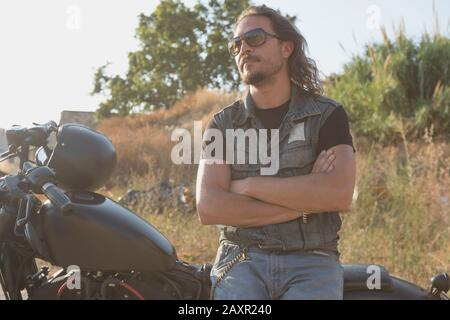 Long-haired brunette guy in sunglasses jeans and a jeans shirt posing on a black custom motorcycle Stock Photo