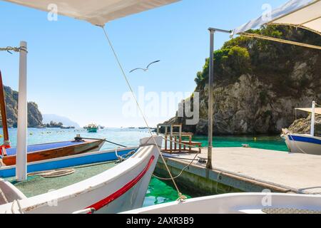 A lone seagull flies past boats at a dock with more boats in the bay at Palaiokastritsa beach on the island of Corfu Greece. Stock Photo