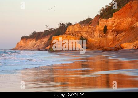 The glowing in the morning light sedimentary rocks on Five Mile Beach at Richardsbay-Meerensee are reflected in the wet sand. Stock Photo