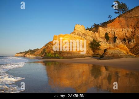 The glowing in the morning light sedimentary rocks on Five Mile Beach at Richardsbay-Meerensee are reflected in the wet sand. Stock Photo
