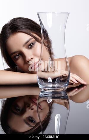 Girl hides her face behind a glass vase. Beauty portrait of young woman at the mirror table. Female on gray background. Stock Photo